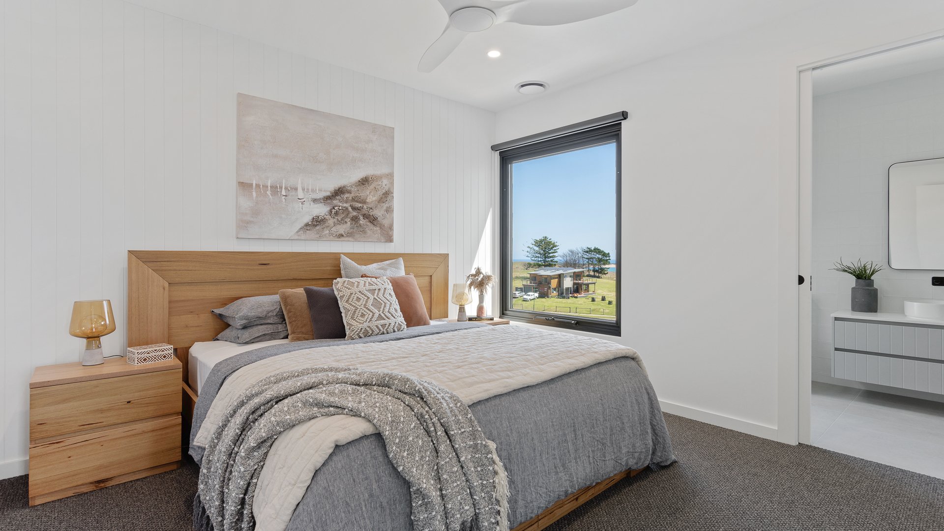 A cozy bedroom with a wooden headboard, decorated with multiple pillows, next to a nightstand with a lamp and decorative item. A large window shows a view of a green landscape with a house and trees. Adjacent to the bedroom is a bathroom with a sink,