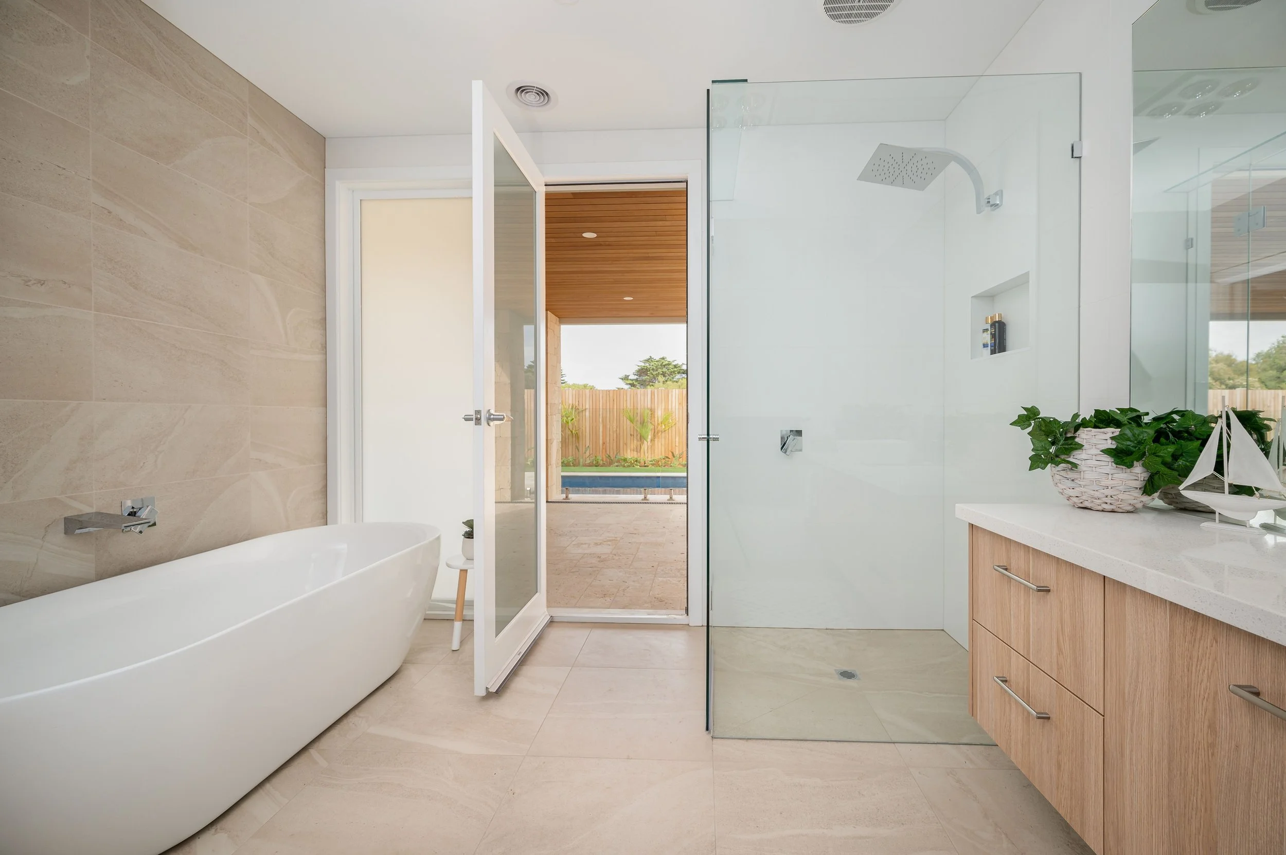 Modern bathroom with a white bathtub, glass shower, wooden cabinet, and view of outdoor pool area.