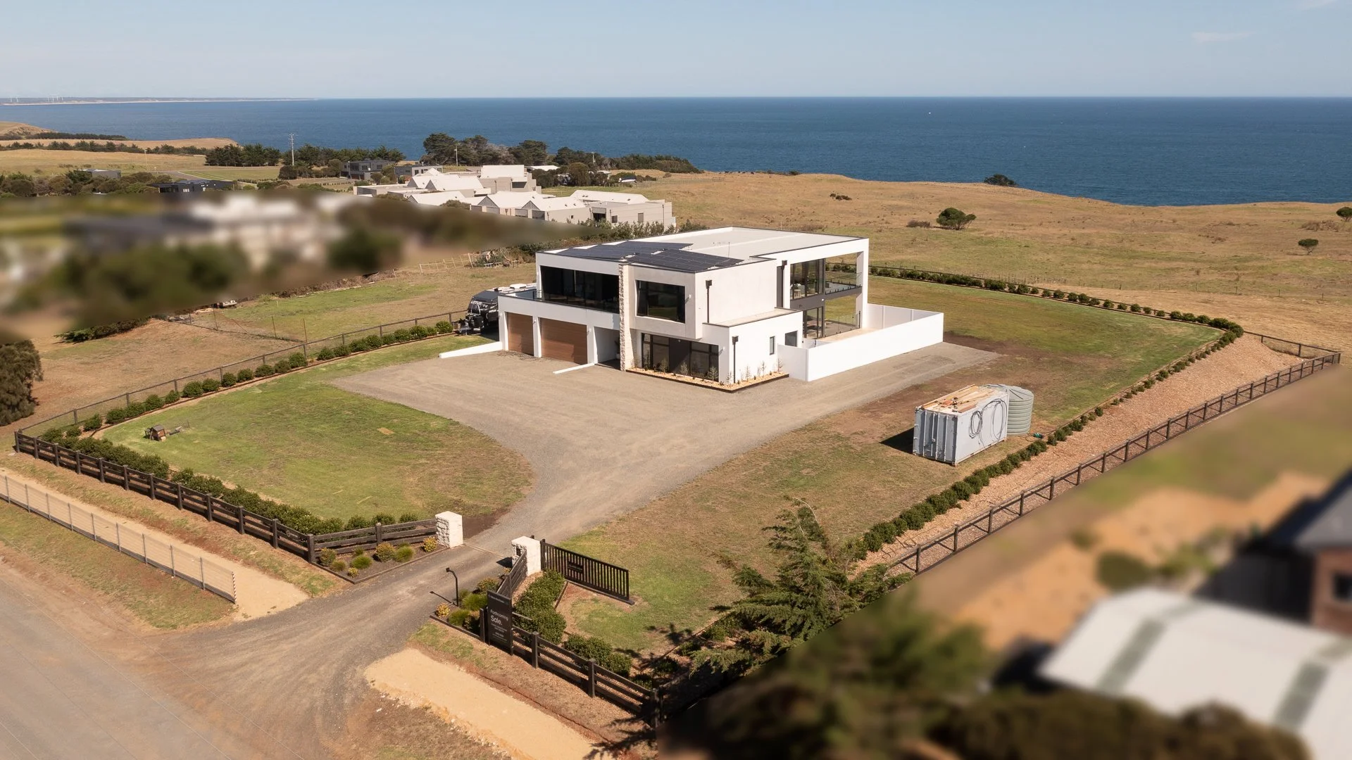 A modern two-story house with white exterior walls, large windows, and a flat roof, situated on a spacious property overlooking the ocean, with a driveway, fenced yard, and minimal landscaping.