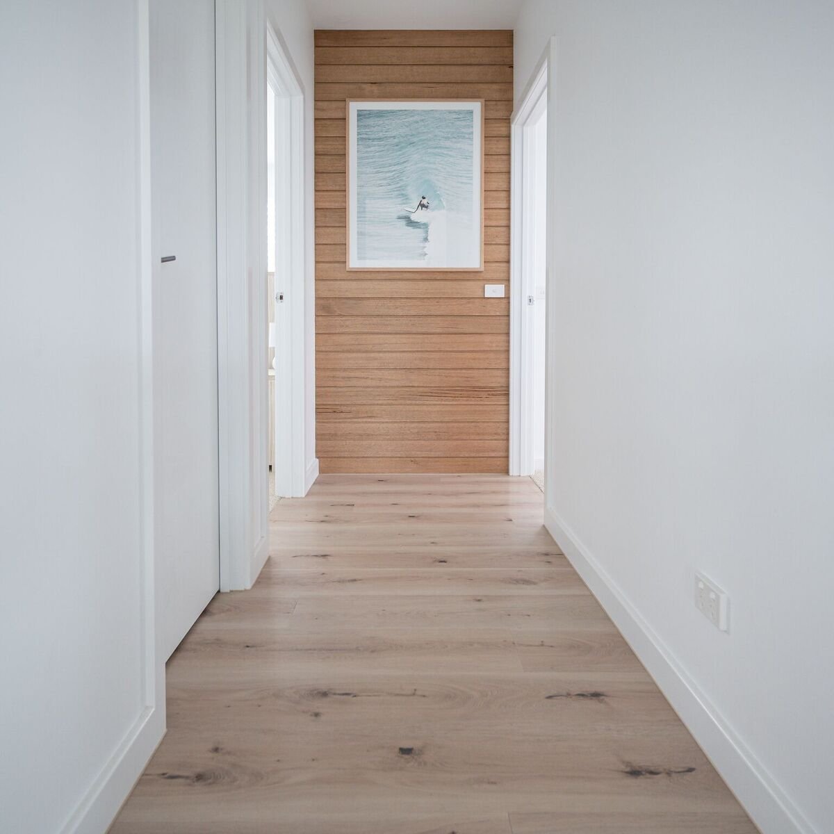 Indoor hallway with light wooden flooring, white walls, and a wooden accent wall at the end featuring a framed ocean painting.