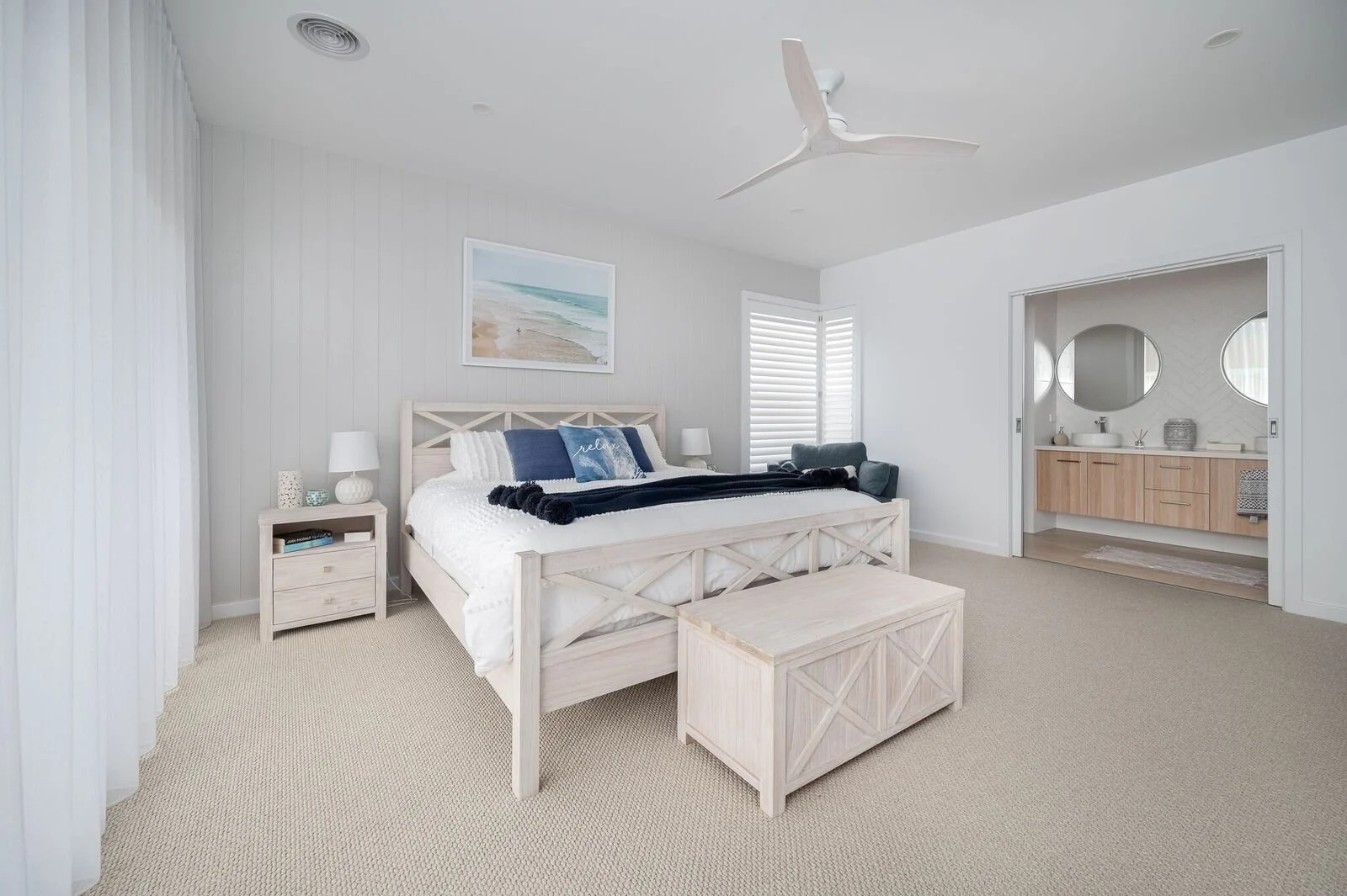 Bright and airy bedroom with a king-sized bed, white wooden furniture, a beige carpet, and an en suite bathroom with round mirrors.