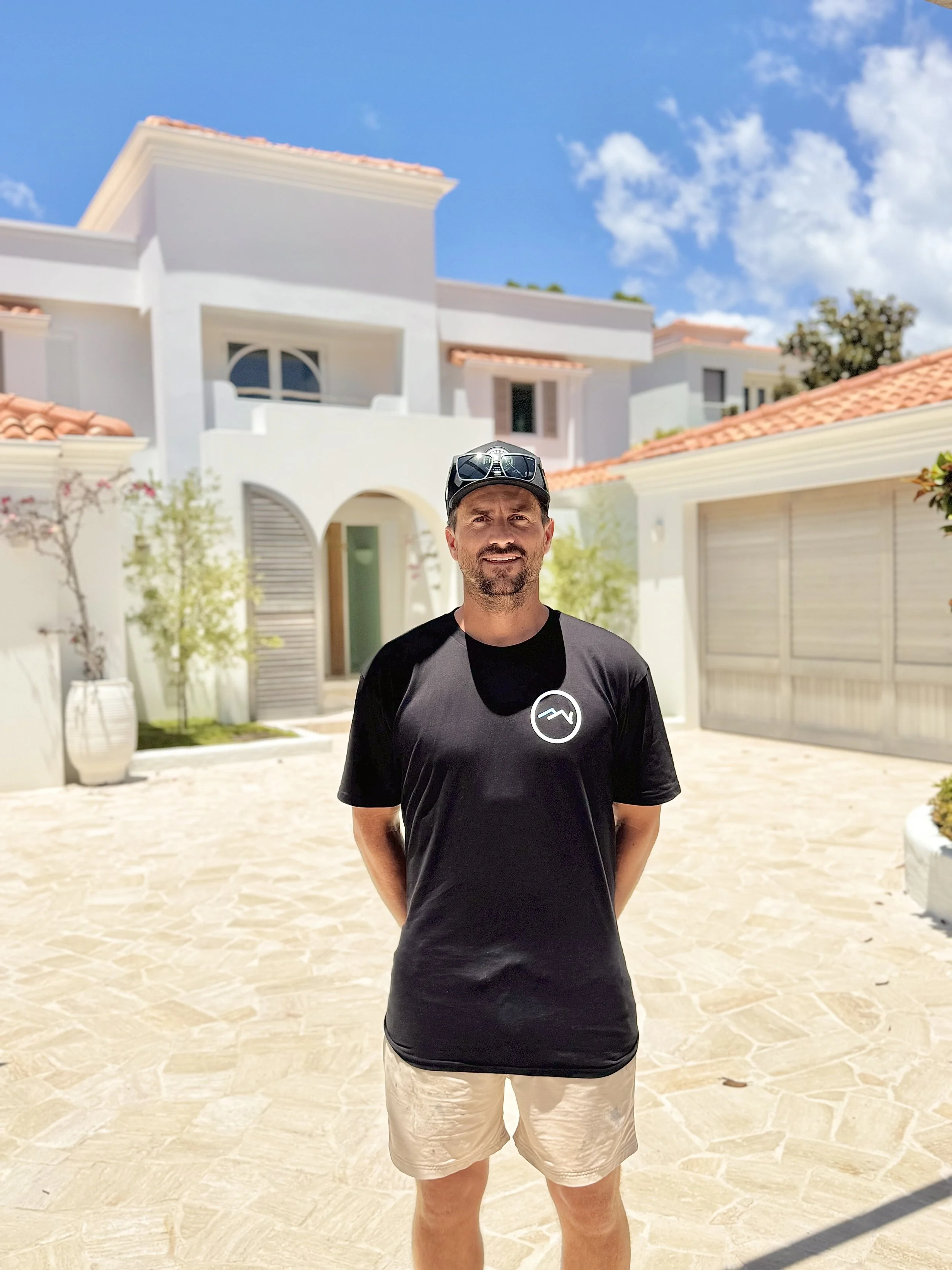 A man standing in front of a modern white house with a red tile roof, blue sky, and some clouds, wearing sunglasses on his cap, a black t-shirt with a white logo, and beige shorts.
