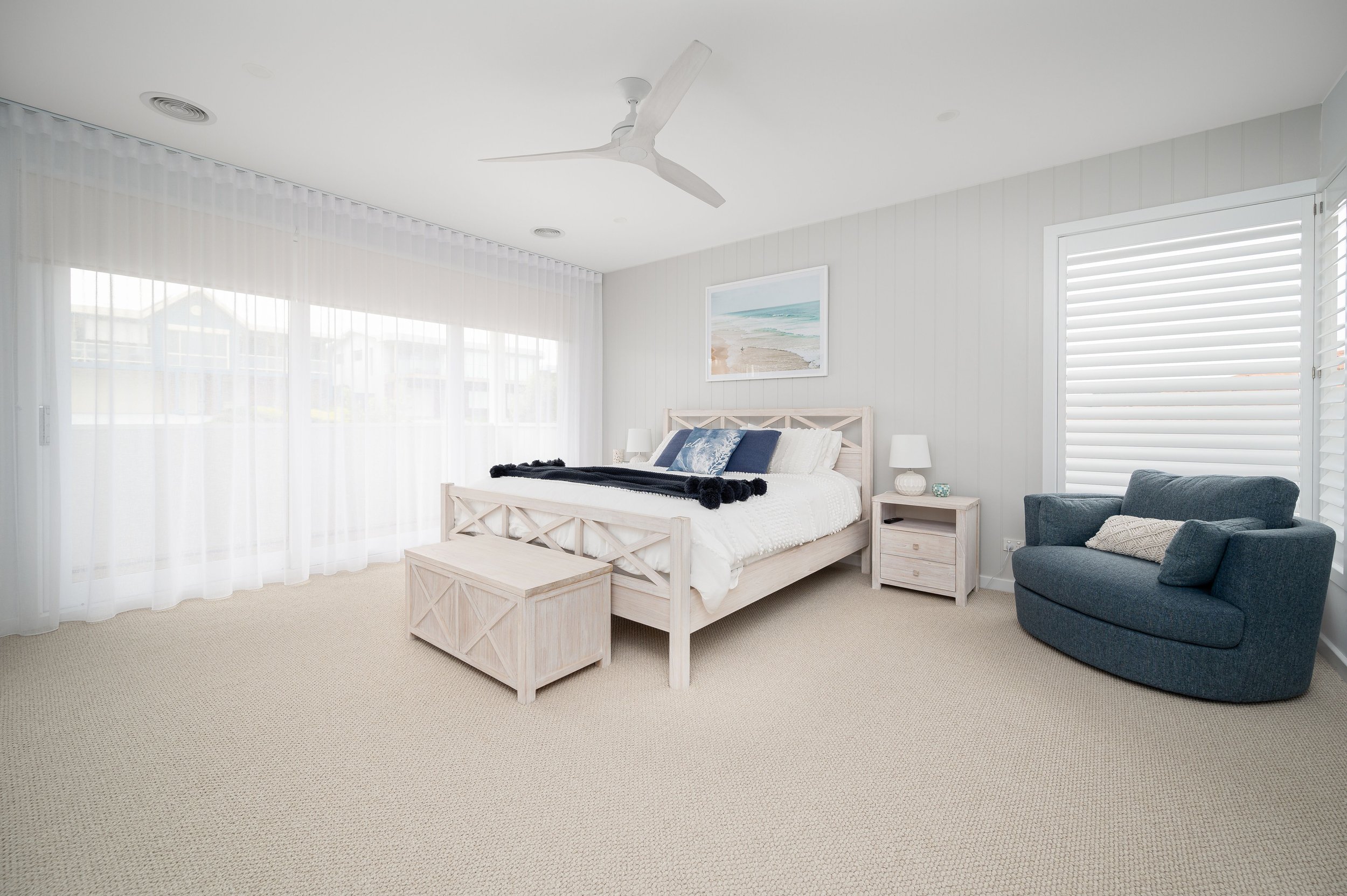 Bright bedroom with white walls and beige carpet, large window with white curtains, bed with white bedding and blue pillows, nightstands with lamps, a blue armchair, and artwork of a beach scene on the wall.