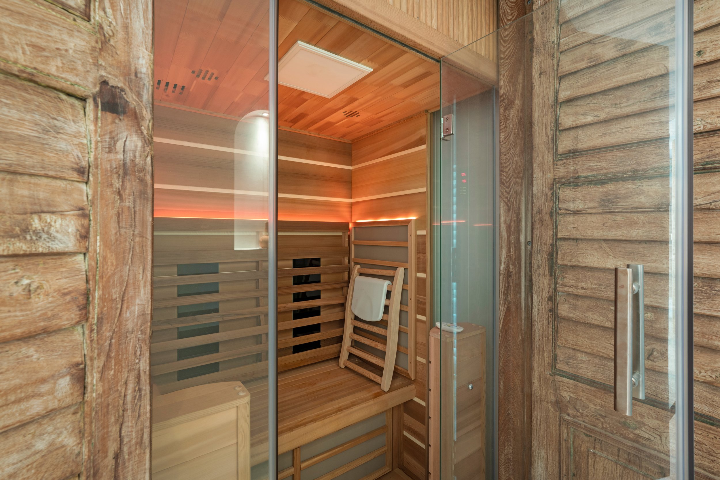 Inside a wooden sauna with a glass door, wooden benches, and a towel hanging on a rack, illuminated by warm lighting.