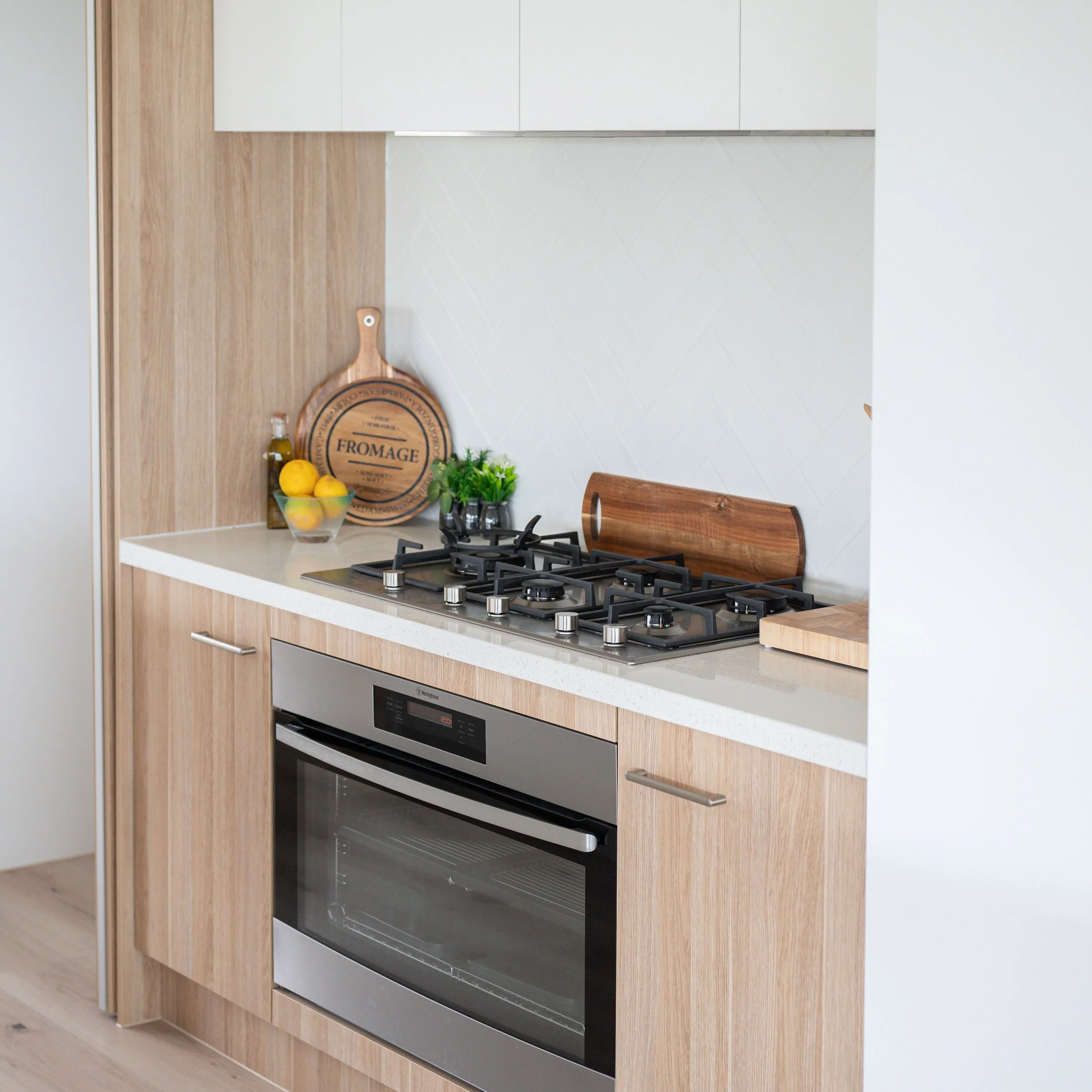 Modern kitchen with built-in oven, gas stove, wooden cabinets, white countertop, and decorative items including a cheese board, lemon bowl, and potted plant.
