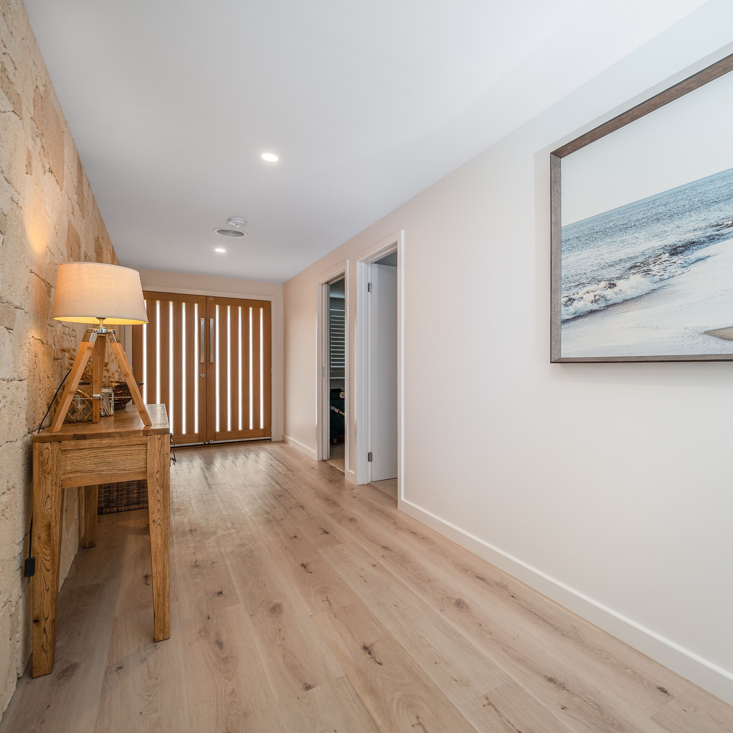 Empty hallway with wooden floor, white walls, a framed beach photo on the wall, a lamp on a side table, and a closed wooden entry door at the end of the hall.