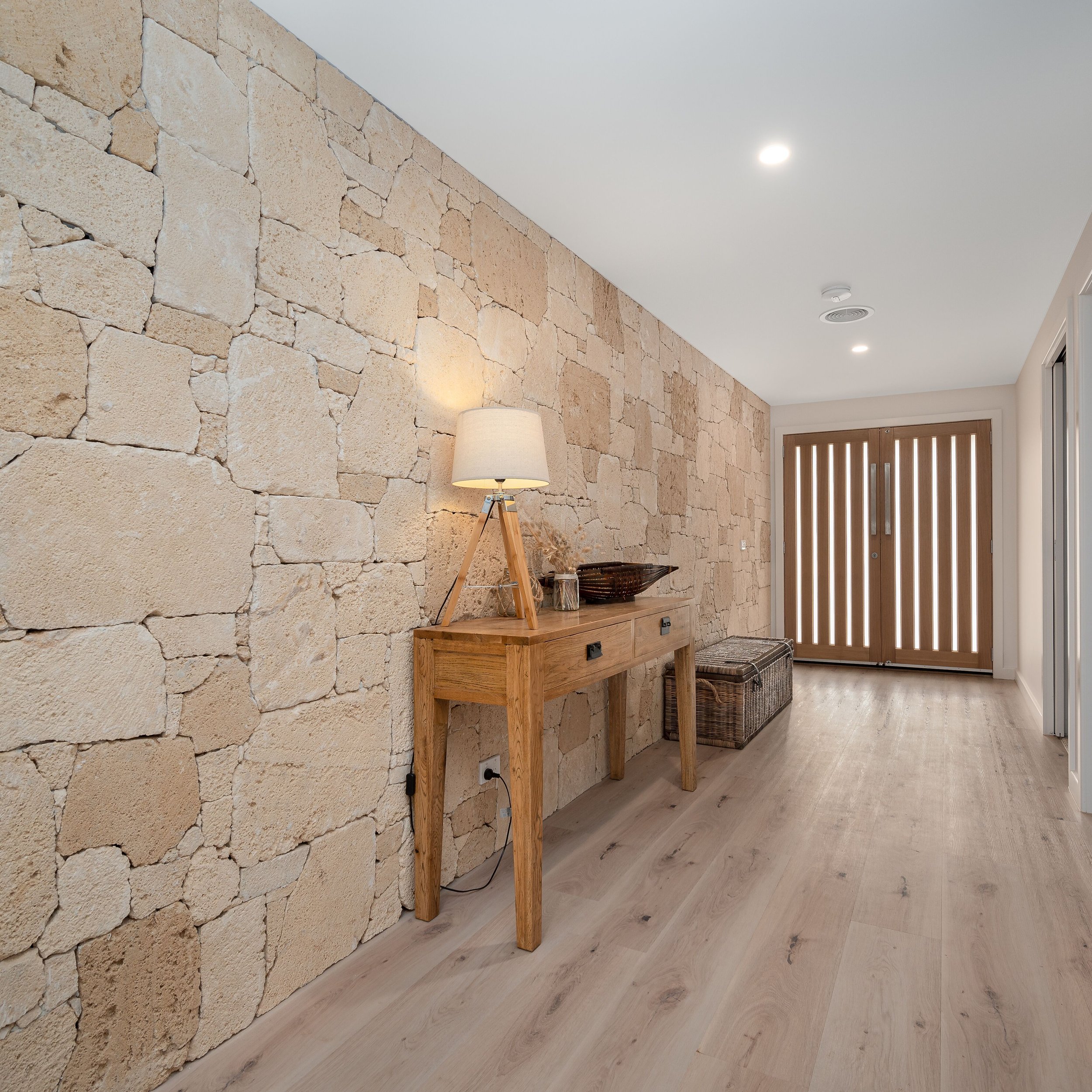 Entryway with stone accent wall, wooden console table with a lamp, basket, and decorative items, wooden front door with striped panels, light hardwood floors, and ceiling lights.