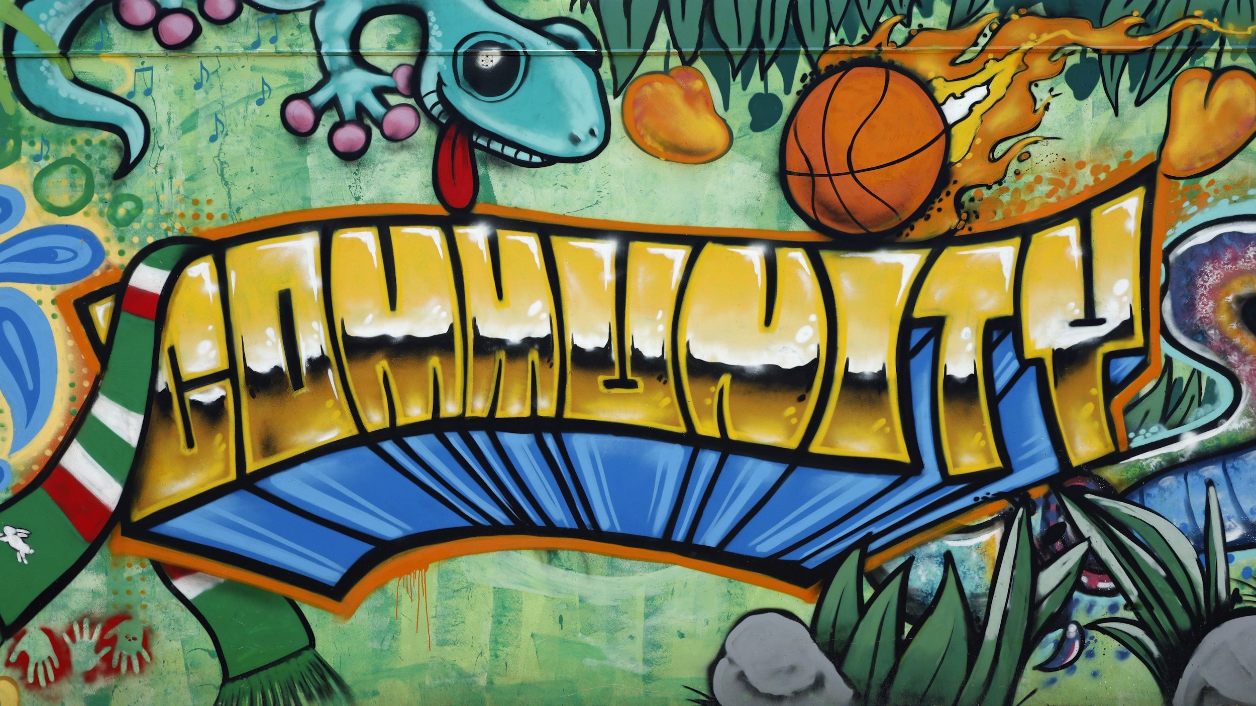 Colorful graffiti mural spelling out 'HUMAN' in bold yellow letters with black outlines, surrounded by vibrant images of a blue gecko, a basketball, and lush green foliage.