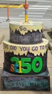 A decorated trash can with yellow, purple, and black paint, featuring inscriptions, and large colorful number and letter cutouts spelling 'STL 150' at the sidewalk.