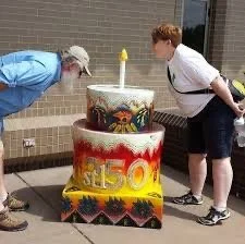 A woman and a boy are smelling a large, decorated cake shaped like a 50th birthday cake outdoors near a building.