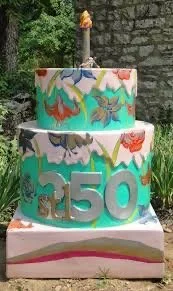 Three-tiered cake decorated with colorful dragon or dinosaur designs and the number '250' on the front, with a birthday candle on top, outdoors in front of a stone wall.