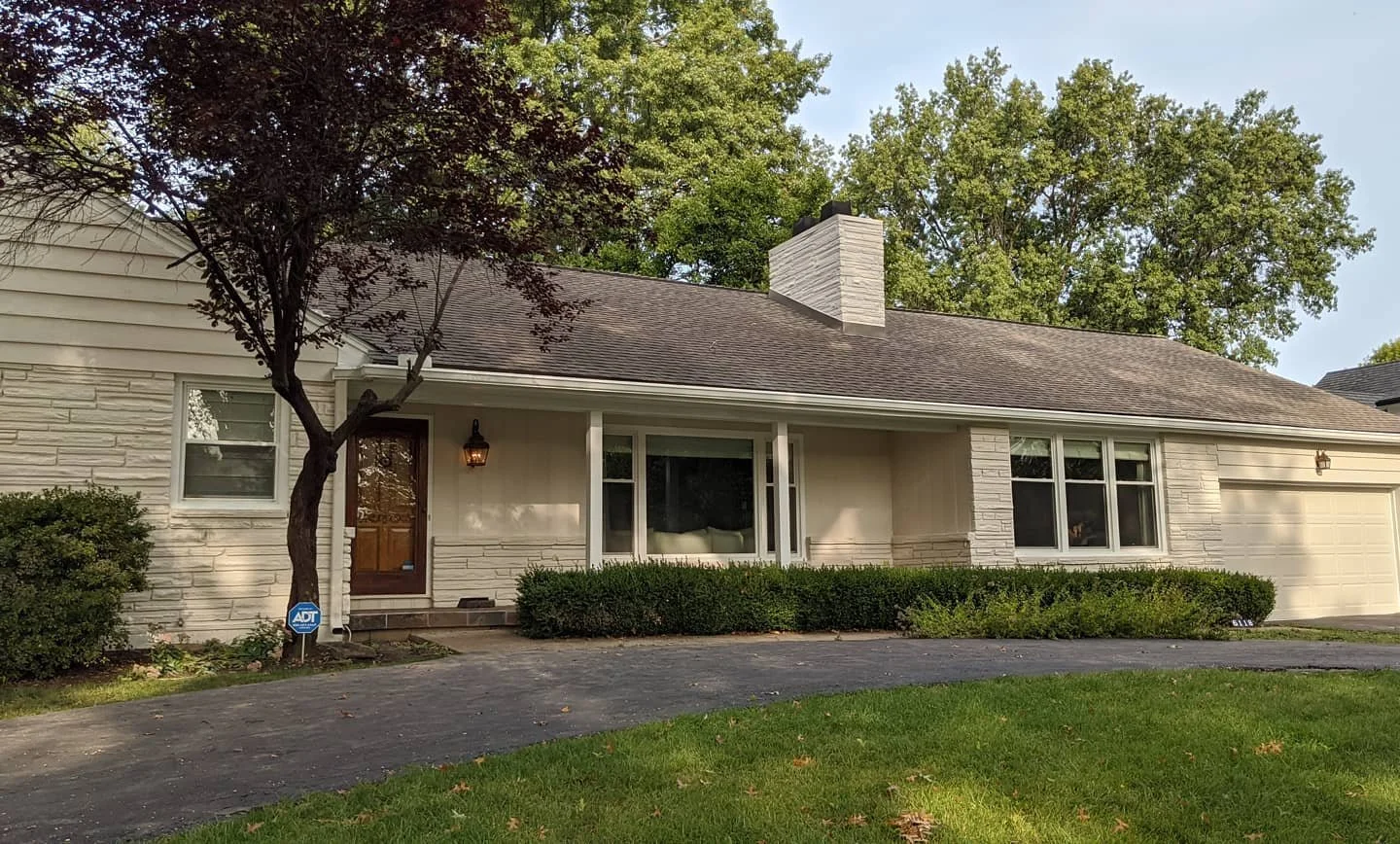 Single-story suburban house with white brick exterior, brown front door, large windows, and attached garage; surrounded by greenery, trees, and a well-maintained lawn.