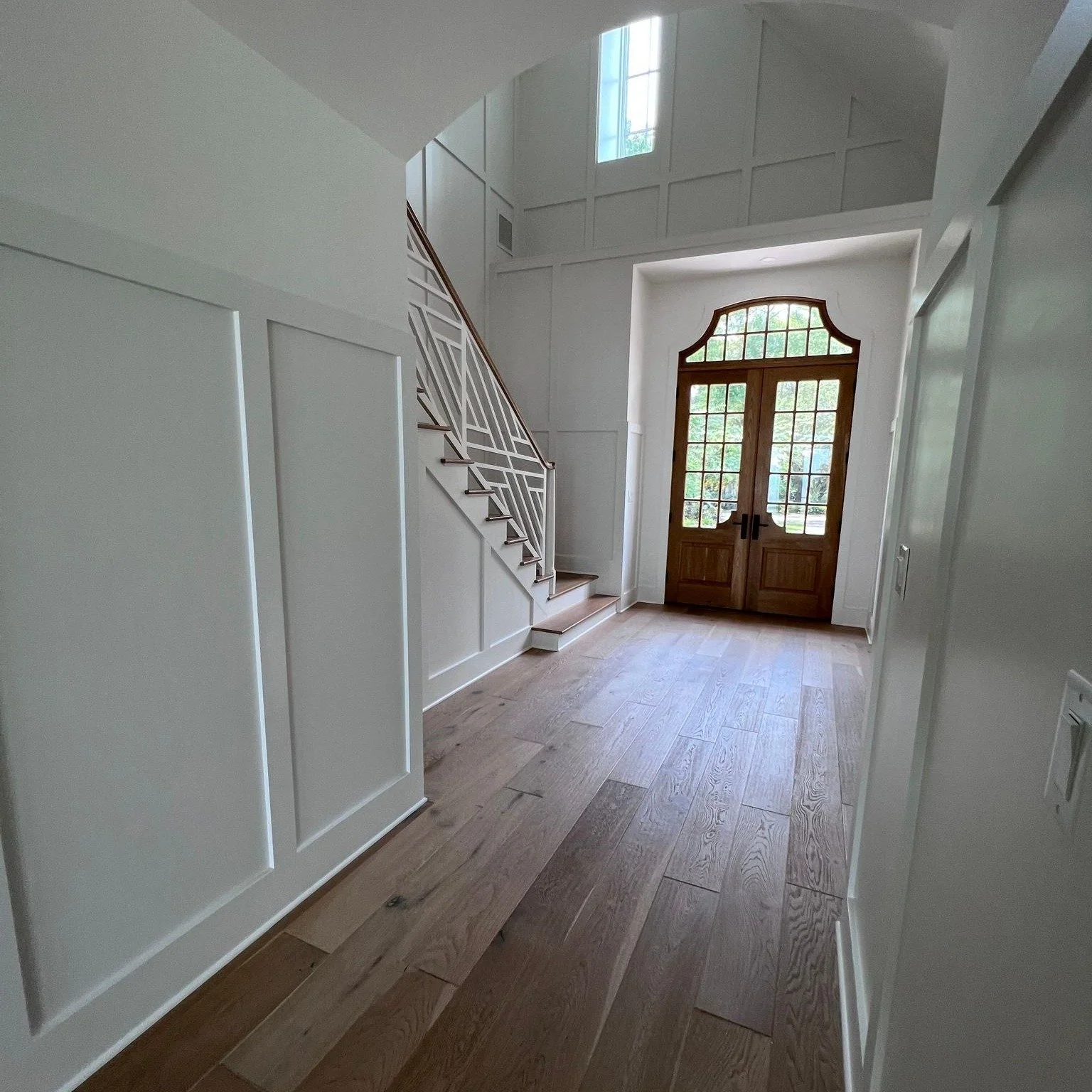 Entryway with wooden front door with glass panels, wooden floors, white paneled walls, staircase with decorative railing, and high ceiling with windows.