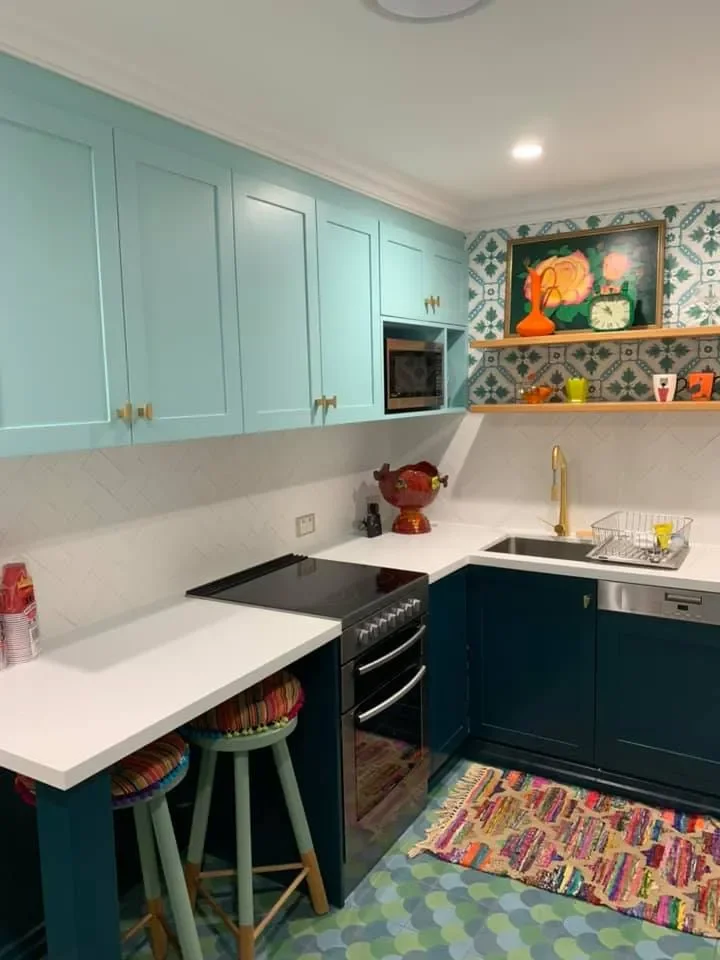 Colorful kitchen with light blue upper cabinets, dark blue lower cabinets, white countertops, and patterned wallpaper. Includes a small dining area with two stools, a stove, microwave, sink with gold faucet, and various decorative items.