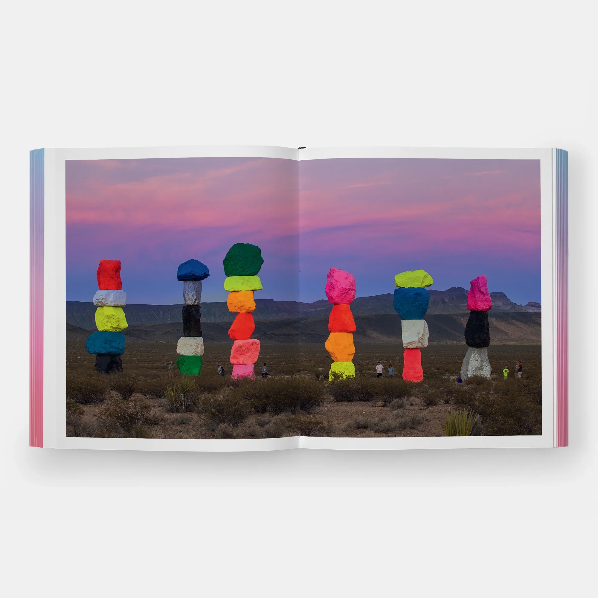Rainbow Dreams Book by Olga Rei and Valentine Uhovski. Phaidon. Colorful stacked rocks installation in a desert landscape during sunset, with pink and purple sky and distant mountains.