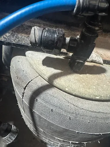 Close-up of a cracked concrete sewer cover with a black pipe fitting attached, and a blue hose nearby.