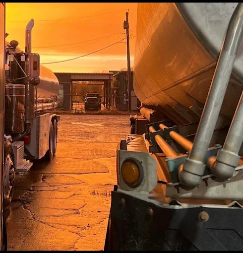 The photo shows a view from a parking lot at sunset, with large trucks and a car in the background. A tanker truck is on the left, and a semi-truck trailer is on the right, with some pipes and equipment mounted on it.