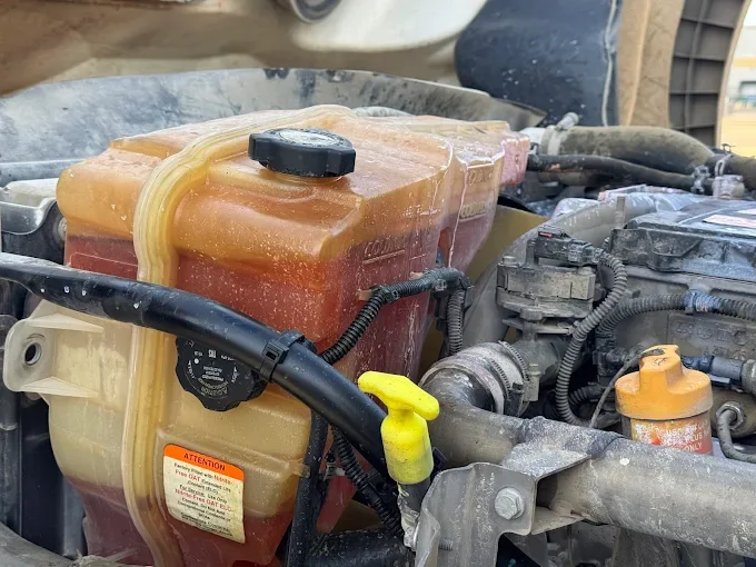Close-up of a car engine compartment showing a large rectangular coolant reservoir made of semi-transparent plastic with orange and yellowish fluid inside. The reservoir has a black cap and a yellow dipstick attached to it. Surrounding components inc