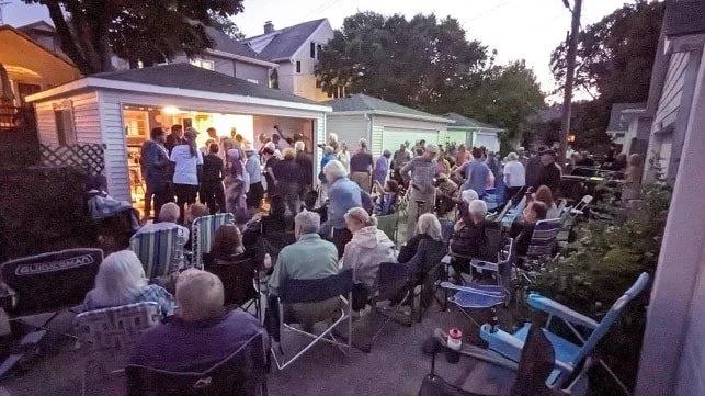 People gathered outside in a backyard during an evening social event, some sitting on chairs and others standing, with a lit indoor area in the background.
