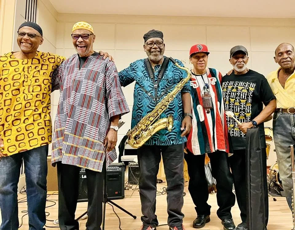 Six men standing together, some holding musical instruments, smiling, in a room with stage equipment.