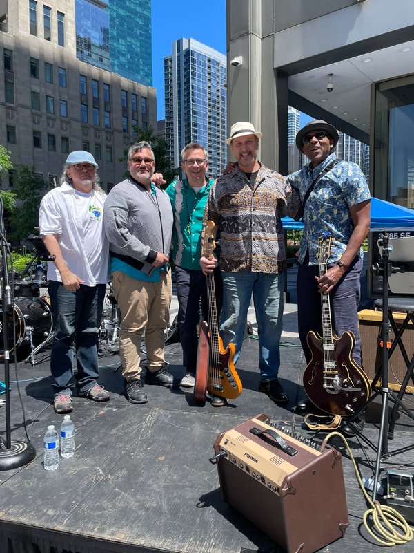 Five musicians standing on an outdoor stage with musical instruments and equipment, city buildings in the background.