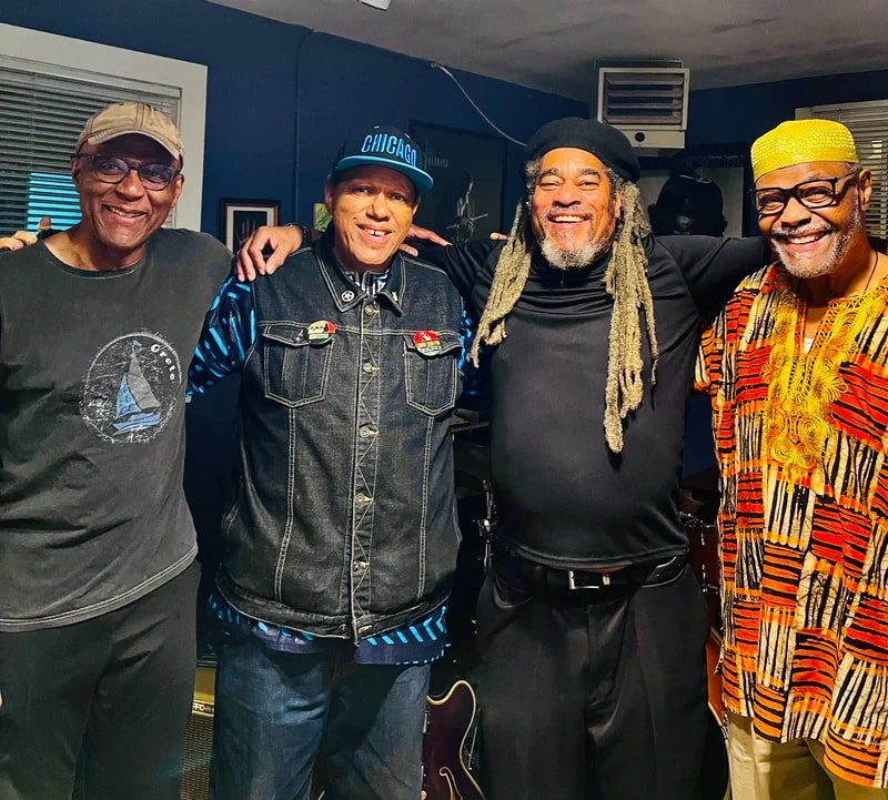 Four men standing together indoors, smiling and posing for the photo. They are casually dressed, with one wearing a colorful African-inspired shirt, another in a black jacket with a blue cap that says 'Chicago', and a third in dark clothing with drea