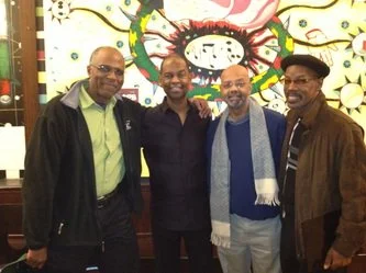 Four men standing together indoors, smiling, with a colorful abstract mural in the background.