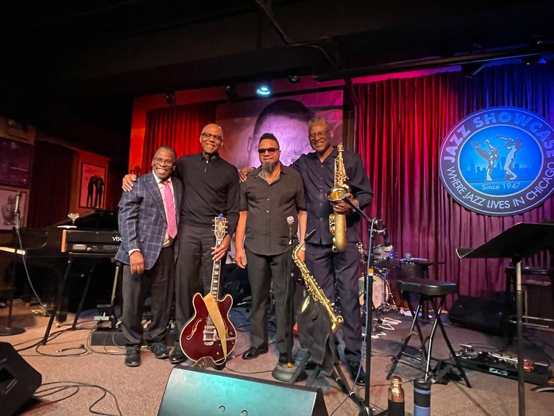 Five jazz musicians standing on stage with instruments, smiling, at the Jazz Showcase in Chicago with a red curtain background and venue sign.