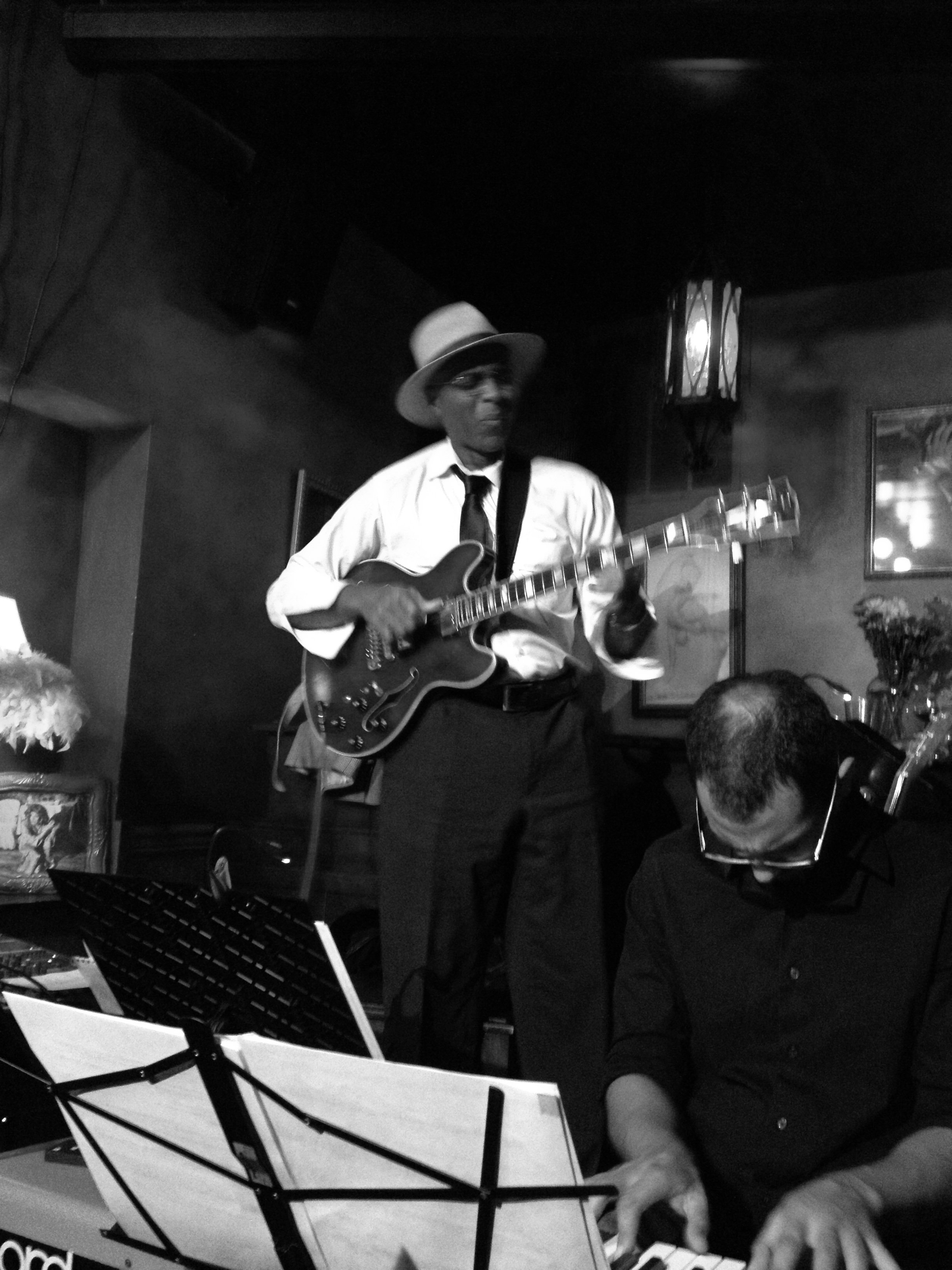 Two musicians performing in a dimly lit venue, with one playing an electric guitar and the other on a keyboard.