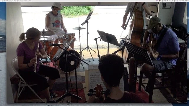 A group of five musicians playing various instruments including drums, clarinet, upright bass, and violin, in an indoor setting near a large window with natural light.