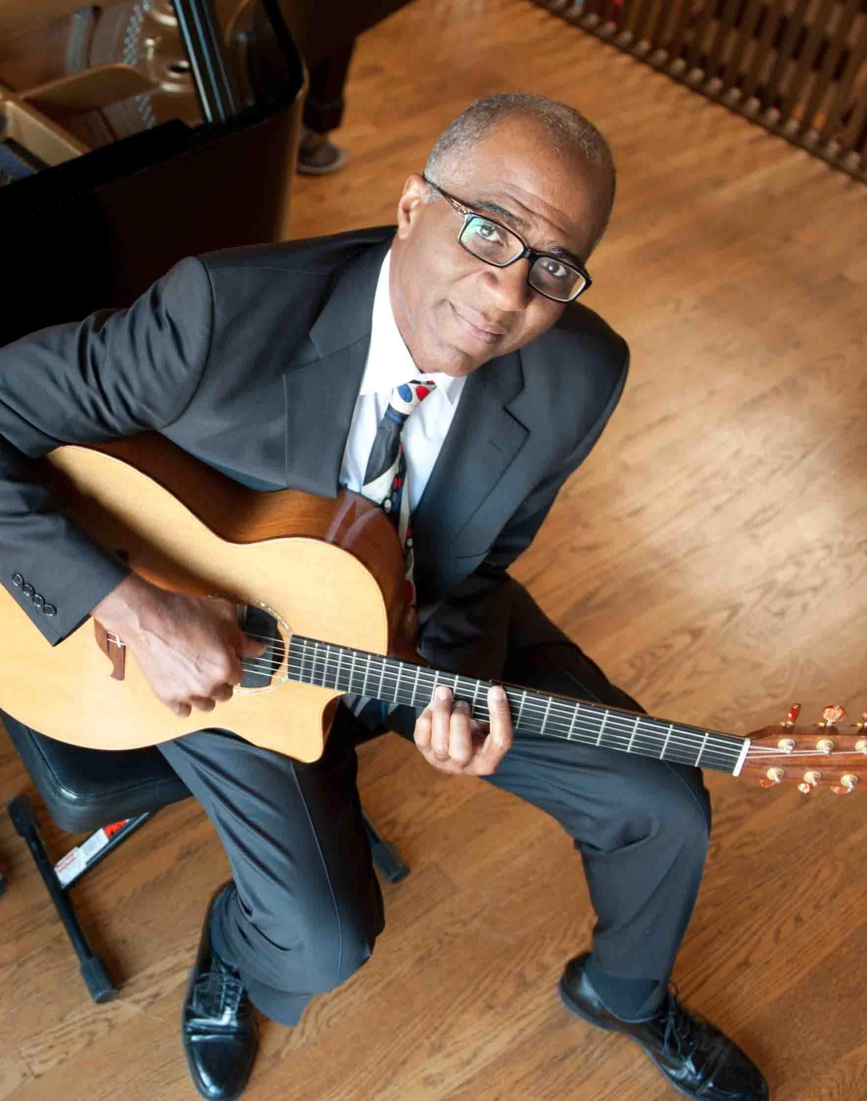 A man in a suit with glasses and a colorful tie sitting on a black chair, holding an acoustic guitar, looking up at the camera, with a wooden floor in the background.