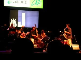 A band performs on stage with musicians playing string instruments and a keyboard, with a singer on the right, under stage lighting.