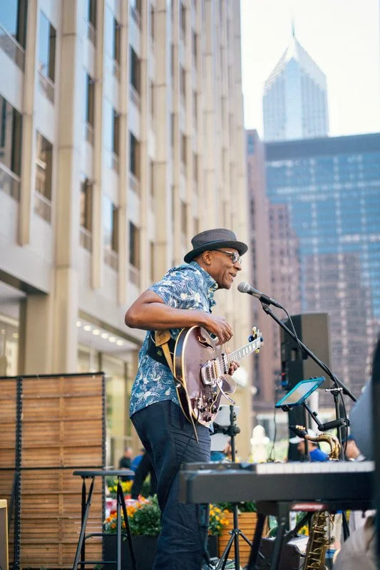 A street musician playing a guitar and singing into a microphone outdoors in a city, with tall buildings in the background, wearing a hat and sunglasses.