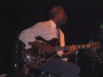 Man playing an electric guitar on stage in a dimly lit setting.