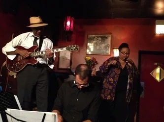 Three musicians performing in a dimly lit room: a man playing guitar, a woman singing, and a seated man in the background.