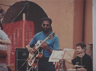 A man with a beard playing an acoustic guitar at an outdoor event, with a music stand and another musician nearby.
