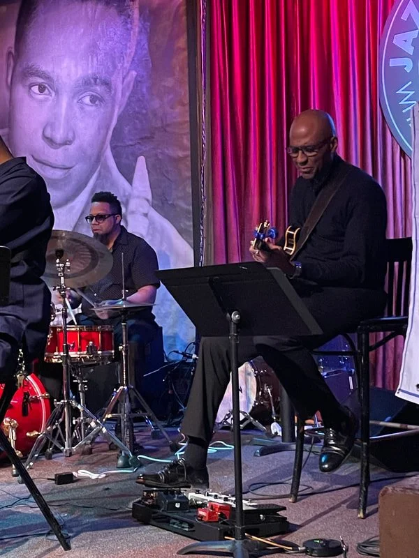 Jazz band performing on stage with a mural of a person's face in the background. The band includes a drummer and a guitarist, with the guitarist seated and playing an electric guitar.