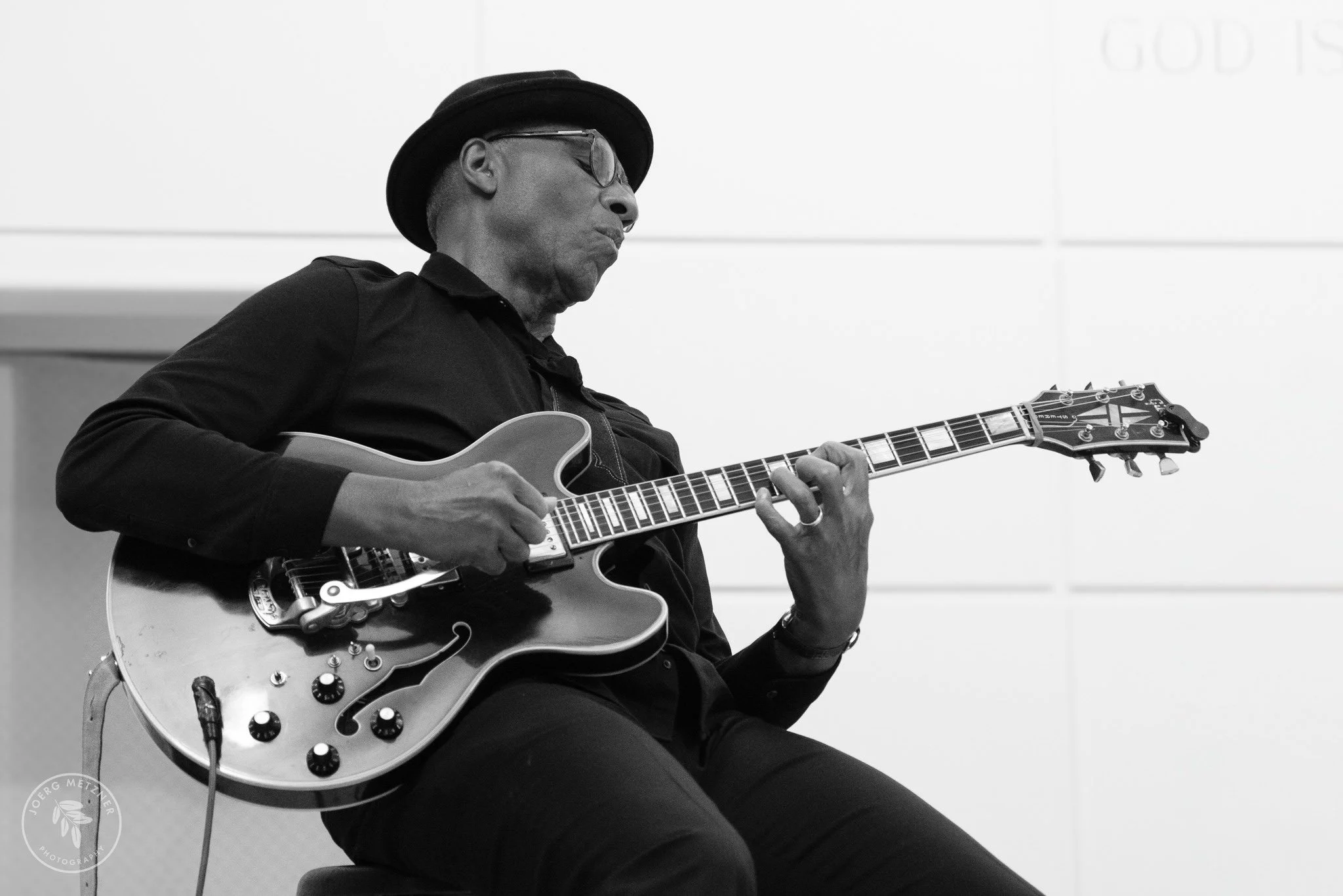 A man wearing a hat and glasses playing an electric guitar in a room with plain walls.