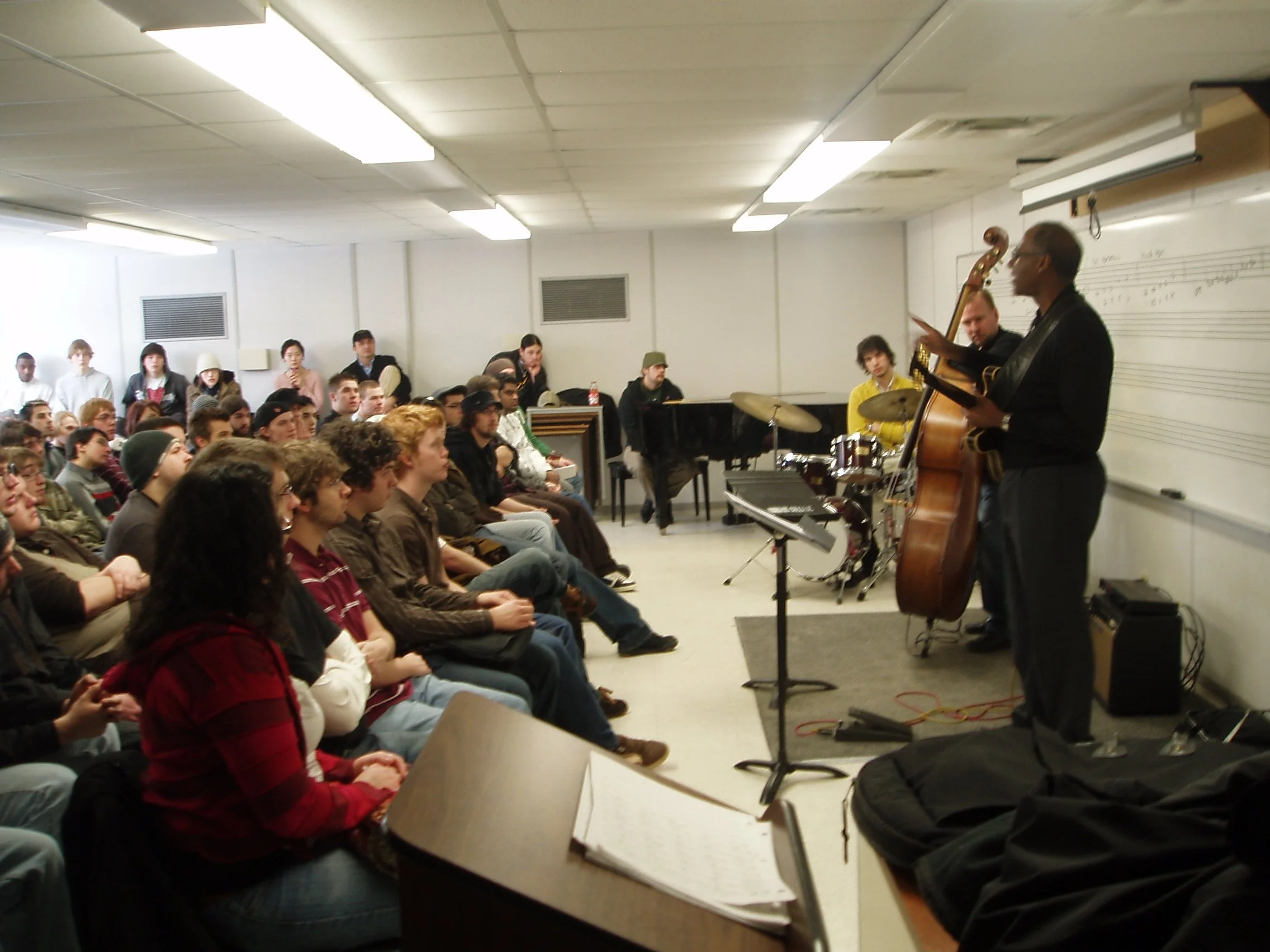 A jazz band performs for an audience in a classroom with white walls and a whiteboard. The band includes a bassist, drummer, and pianist, while the audience consists mostly of young adults seated and watching attentively.