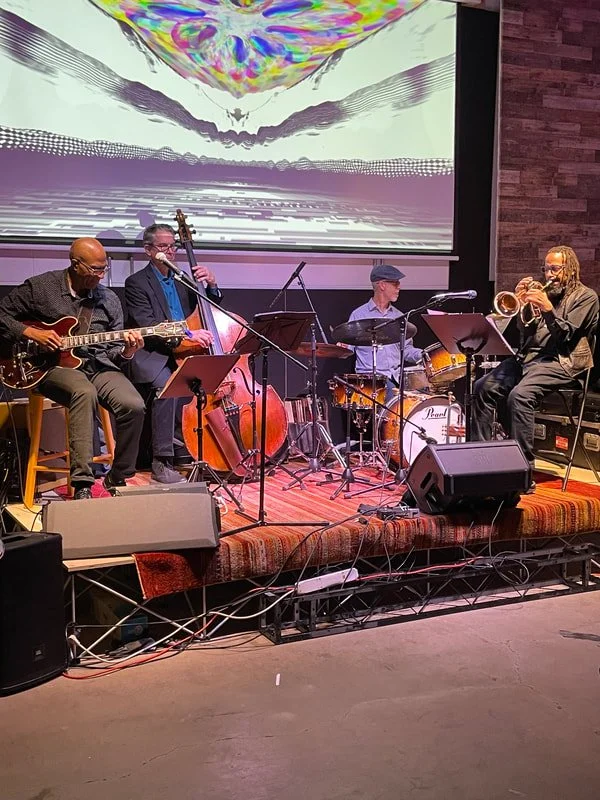 A jazz band performing on stage with four musicians playing guitar, double bass, drums, and trumpet, with abstract digital art projected on a screen behind them.