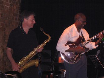 Two musicians performing on stage: one playing a saxophone, the other playing an electric guitar.