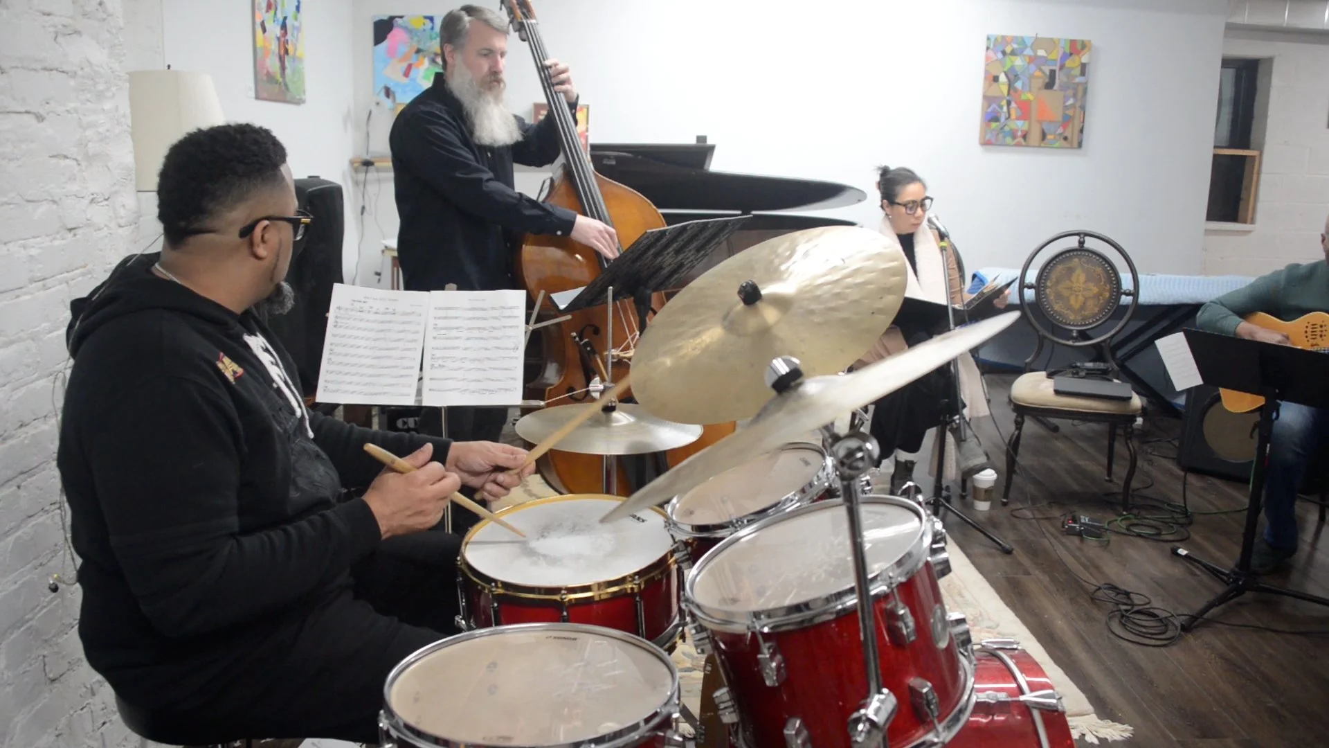 A jazz band performing indoors with a drummer, a double bassist, a pianist, a vocalist, and a guitarist. They are playing in a room with white walls decorated with colorful abstract art.