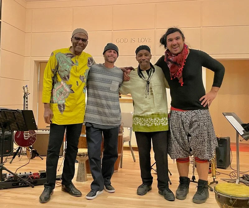 Four men standing together inside a room, smiling. Behind them, a sign reads 'GOD IS LOVE'. Musical instruments and equipment are scattered around, indicating a performance or practice setting.