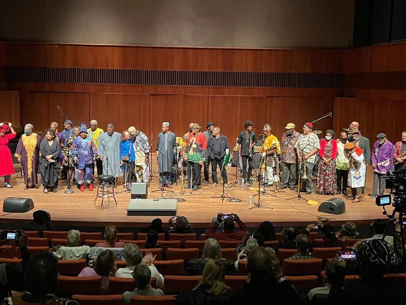 A diverse group of performers standing on stage after a performance, with audience members seated below taking photos.
