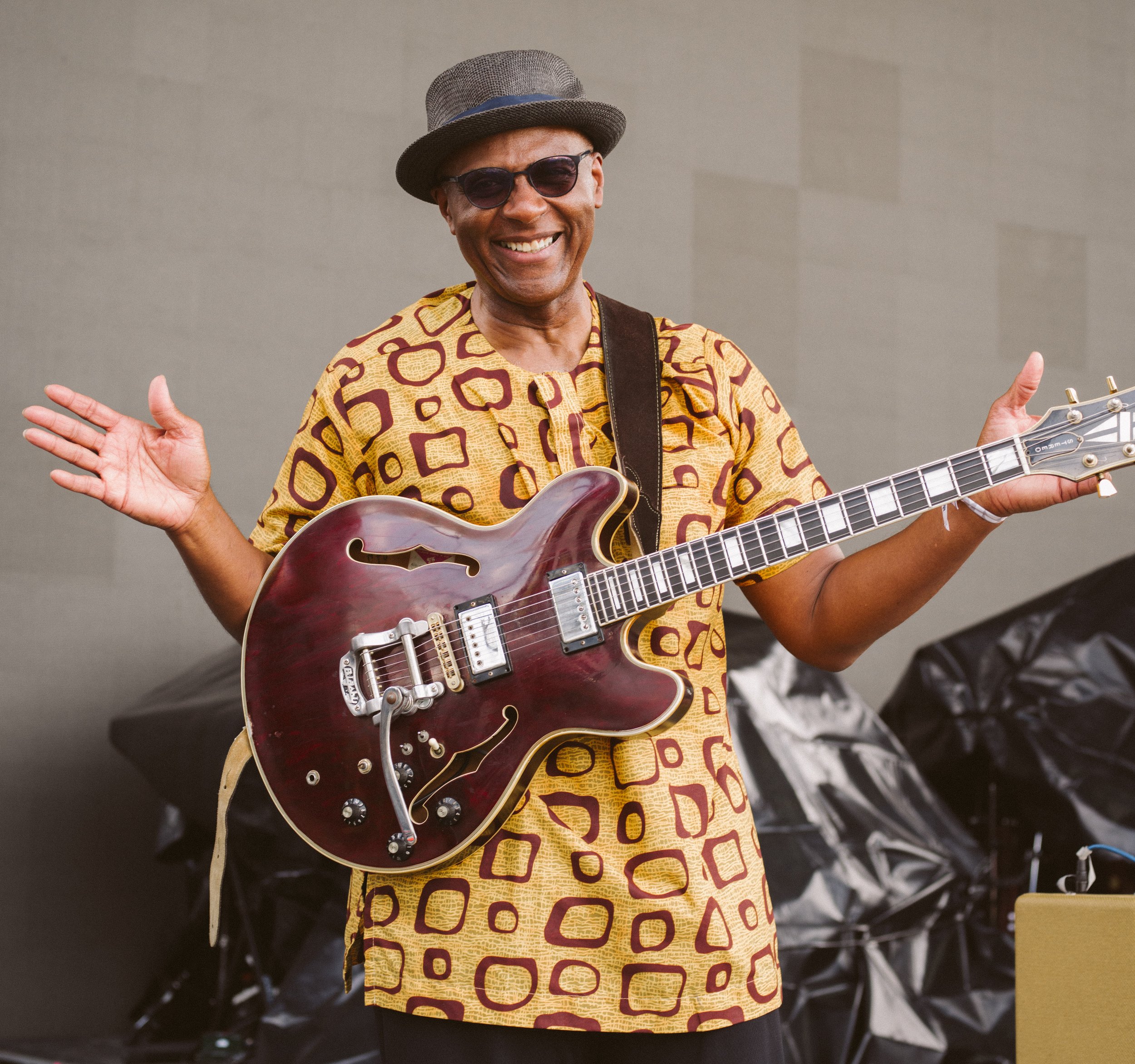 A smiling man wearing a patterned yellow and brown shirt, sunglasses, and a hat, holding a red hollow-body electric guitar and waving.
