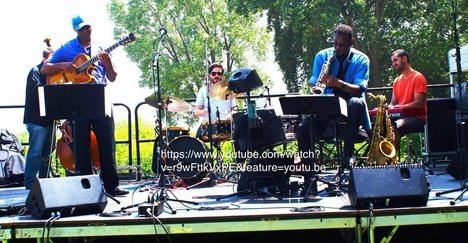 A five-piece band performing outdoors on a stage with trees in the background. The band includes a guitarist, a bassist, a drummer, a keyboardist, and a saxophonist.