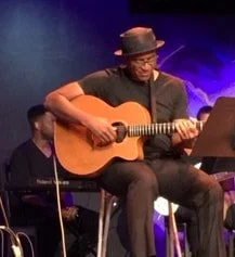 A man playing an acoustic guitar on stage, wearing a hat and glasses, with a dark background and stage lighting.