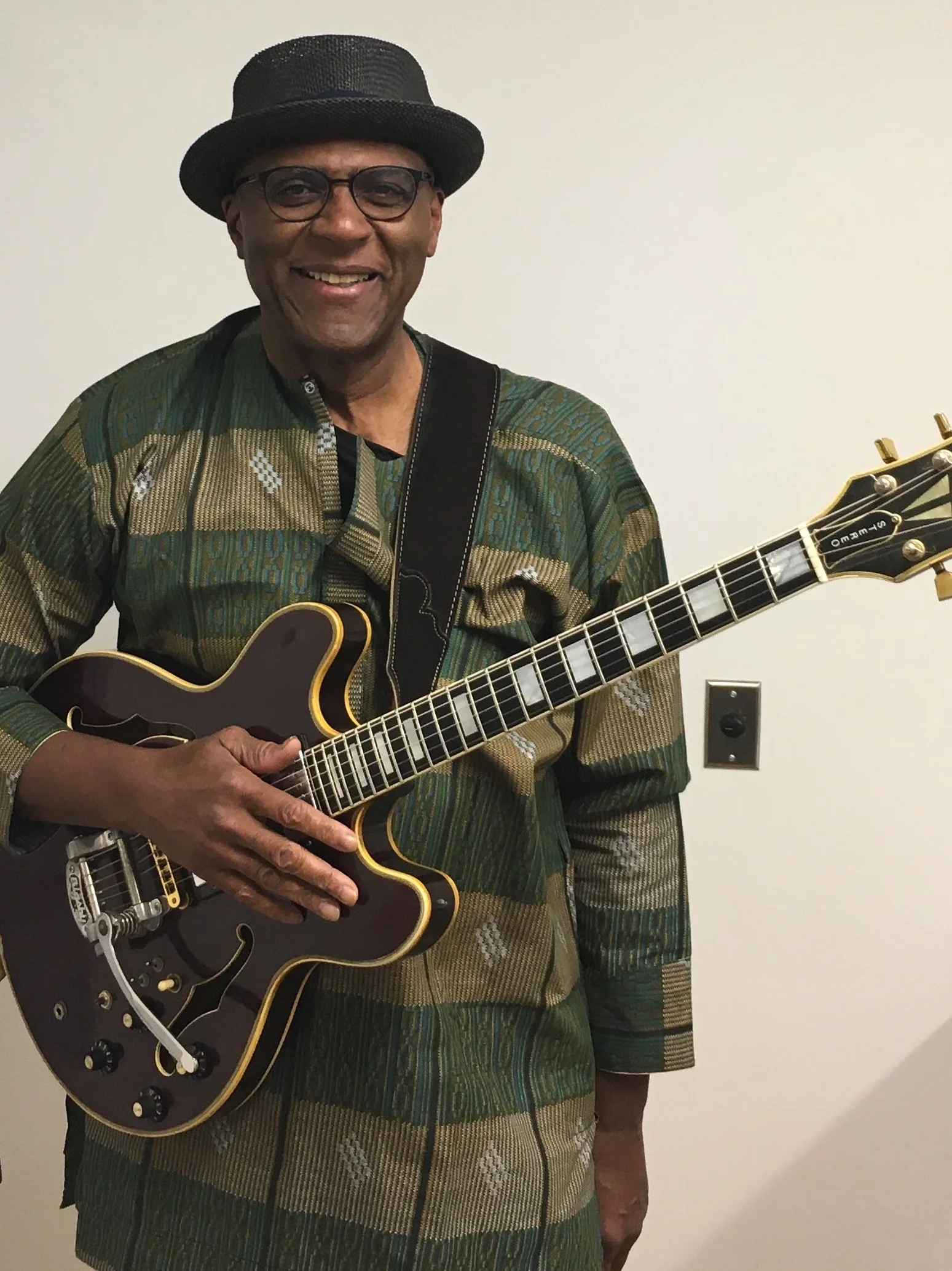 A man wearing glasses, a black hat, and a patterned jacket, smiling while holding a semi-hollow electric guitar against a plain light-colored wall.