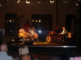 Musicians performing on stage with a guitarist, bassist, and drummer, in a dimly lit venue.