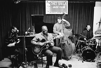 Four musicians performing on stage at the Chet Baker Jazz Club, including a guitarist, a double bassist, a drummer, and a keyboard player.
